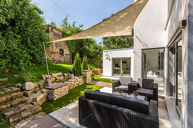 Kategorie Komplettsets Dreieckiges, beiges Sonnensegel im Garten über Terrasse im Sonnenschein mit einem Pfosten und zwei Wandhalterungen über einer schwarzen Sitzgruppe